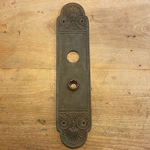May include: A vintage, ornate, dark metal door plate with a circular cutout and a smaller circular cutout below it. The plate has a decorative design around the edges.