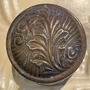 May include: A round, brass door knob with a detailed floral design. The knob has a worn, aged patina.