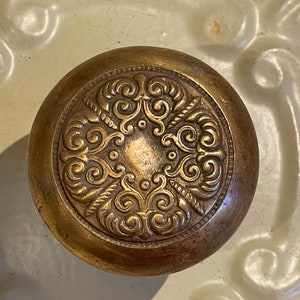 May include: A vintage brass door knob with an ornate, intricate design. The knob is round and has a raised, detailed pattern of swirls and scrolls.