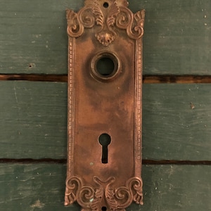 May include: Antique brass door plate with ornate floral designs and a keyhole. The plate has a worn, aged look.
