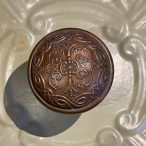 May include: A round, copper-colored door knob with an intricate, ornate design. The knob has a raised, floral pattern with a central, circular motif.
