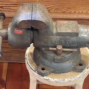 May include: A heavy-duty Wilton bench vise with a weathered gray finish, featuring a prominent metal handle, a swivel base, and a red logo plate labeled 410. The cast iron tool displays the embossed text Wilton and Made in USA on its side.