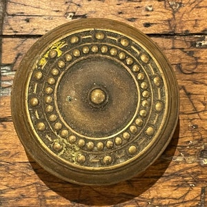 May include: A round, brass-colored door knob with a decorative pattern of small circles around the edge. The knob has a slightly raised center with a small, round knob in the middle.