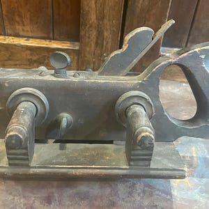May include: Antique wooden hand plane with a dark finish. The tool features a handle, adjustable knobs, and a blade. This woodworking tool is designed for shaping and smoothing wood surfaces. The plane is likely from the early 20th century.