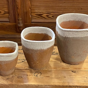 May include: Three terracotta-colored plant pots with a white band near the rim. The pots are different sizes and have a rustic, textured appearance. They are arranged on a wooden surface against a wooden background.