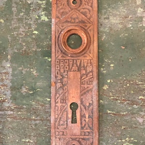 May include: Antique brass door plate with intricate floral and geometric designs. The plate has a keyhole and a round hole for a doorknob.