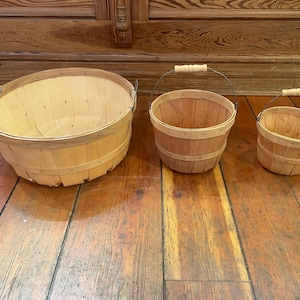 May include: Three wooden baskets of varying sizes. The largest is oval-shaped without a handle, while the other two have wire handles with wooden grips. The baskets are made of light-colored wood and are set on a wooden floor.