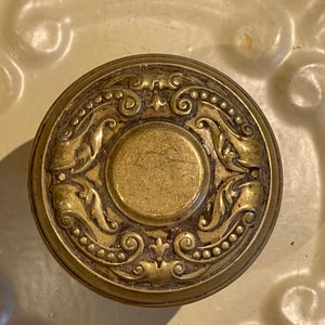 May include: A round, brass door knob with an ornate design featuring a central circle surrounded by intricate scrollwork and floral motifs.