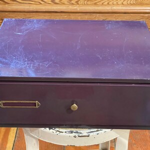 May include: A single drawer metal file cabinet with a purple finish. The drawer has a brass pull handle and a brass label holder.