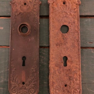 May include: Two antique door plates with ornate designs. The plate on the left is dark brown with a keyhole and a round knob hole. The plate on the right is rusty brown with a keyhole and a round knob hole.
