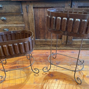 May include: Two brown metal plant stands with oval-shaped planters. The stands are made of wrought iron with a decorative scroll design. The planters are made of copper and have a hammered texture.