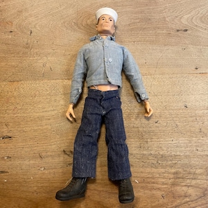 May include: A posable action figure wearing a white sailor hat, a light blue button-up jacket, dark blue denim trousers, and black boots. The figure has a fair complexion and is dressed in a nautical-inspired outfit with visible stitching details on the jacket and pants.