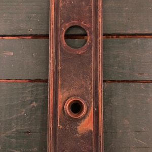 May include: Antique brass door plate with two round holes, one larger than the other. The plate has a worn, aged look with a dark brown patina.