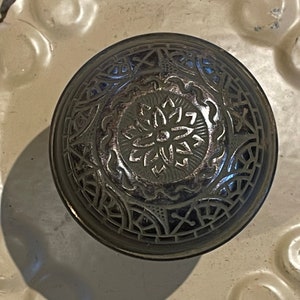 May include: A round, black metal door knob with an intricate, silver-colored design. The design features a central flower motif surrounded by a geometric pattern.
