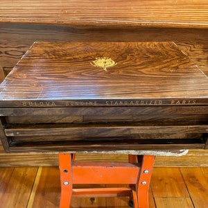 May include: A wooden box with two drawers labeled "BULOVA GENUINE STANDARDIZED PARTS". The box has a gold-colored emblem on the top.