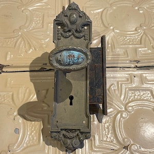 May include: Antique brass door knob and lock set with ornate details. The knob has a decorative oval design and the lock plate has a keyhole and a decorative border.
