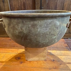 May include: Large, antique bronze cauldron with two side handles. The cauldron has a rounded bowl shape and sits on a wooden stand. The metal has a weathered appearance, suggesting age and use. The background features wooden furniture.