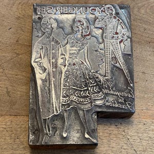 May include: A vintage metal printing plate with the text "Younger Set" and the phrase "Everywhere in Fashion" at the bottom. The plate features three figures in relief, two women and one man, all wearing clothing.