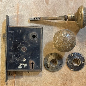 May include: Antique brass door hardware set with a black metal door plate, two round door knobs, and a spindle. The door plate has a keyhole and a square hole. The door knobs have intricate designs.