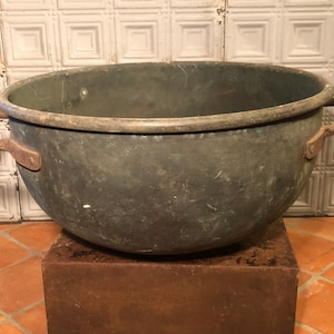 May include: A large, antique metal cauldron with two sturdy handles. The cauldron has a rounded shape and a dark, weathered finish, suggesting age and use. It sits on a square, rusty metal base, adding to its rustic charm. The handles are also rusty.