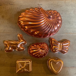 May include: A collection of six copper-colored metal baking molds in various shapes, including a large nautilus shell, a rabbit, a smaller shell, a butterfly, a square mold with a bird design, and a heart-shaped mold, all featuring a metallic finish.