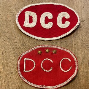 May include: Two red oval patches with white stitched borders. The top patch has the letters "DCC" in white stitching. The bottom patch has the letters "DCC" and three gold stars.