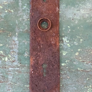 May include: A rusty metal door plate with an ornate design and a keyhole. The plate is rectangular and has a circular hole in the center.
