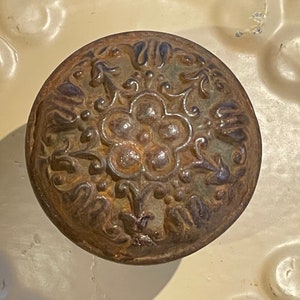 May include: A round, ornate, metal knob with a floral design. The knob is rusty and has a dark brown color.