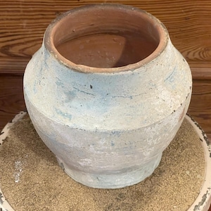 May include: A wide-mouthed terracotta vessel with a weathered, pale blue and white painted exterior finish. The pot features a rounded body that tapers toward the base and a wide, open circular rim, revealing the natural reddish-brown clay interior.