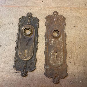 May include: Two antique door plates with ornate, floral designs. One is a tarnished brass color, the other is heavily rusted. Both have keyholes and circular openings for a door knob.
