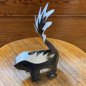May include: A metal sculpture of a skunk with a black and white body and a tall, stylized tail. The sculpture is made of metal and sits on a wooden table. The skunk's tail is made of white and black metal.