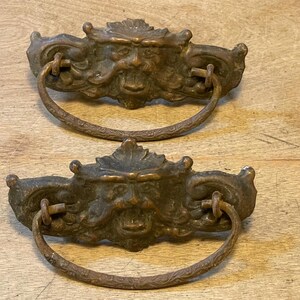 May include: Two antique bronze drawer pulls. Each pull features a detailed face design with a ring handle. The bronze has a weathered appearance, suggesting age and use. The pulls are likely for a cabinet or drawer.