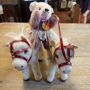May include: Two white plush toy horses with red leather bridles and a white plush teddy bear wearing a pink dress and holding a purple flower.
