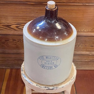 May include: A cylindrical stoneware jug with a glossy dark brown glazed top and a matte off-white base. The front features a blue circular stamp reading THE WESTERN POTTERY CO. MADE IN DENVER. A small cork stopper is inserted into the narrow neck.