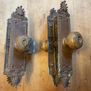 May include: Two antique brass door knobs with ornate detailing and keyholes. The knobs are mounted on a wooden surface.