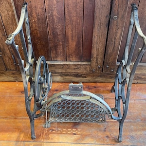 May include: Antique cast iron sewing machine base with ornate scrollwork and a decorative wheel. The base has a weathered, aged appearance, with a rectangular basket-like structure in the center. The legs are curved and feature decorative cutouts.