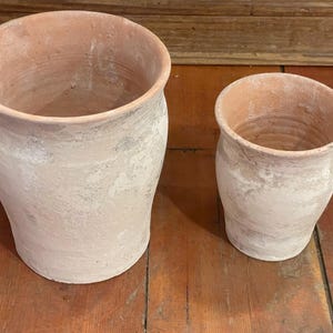 May include: Two terracotta pots with a weathered, matte finish in shades of light tan and dusty white. The pots feature a wide, open circular rim that tapers down to a narrower base. The surface texture appears porous and uneven throughout.