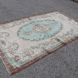 Könnte beinhalten: Ein Vintage-Teppich mit einem hellbraunen Rand und einem cremefarbenen Hintergrund. Der Teppich hat ein großes, blaugrünes Blumenmuster in der Mitte und kleinere Blumenmuster am Rand.