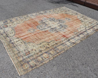 Op de afbeelding: Een vintage tapijt met een vervaagde oranje, blauwe en crème kleurstelling. Het tapijt heeft een bloemenpatroon en een verweerde uitstraling.
