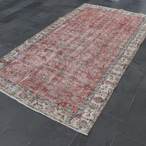 Könnte beinhalten: Ein rechteckiger, abgenutzter Teppich mit verblasstem roten und cremefarbenen Blumenmuster. Der Teppich hat einen cremefarbenen Rand mit floralen und geometrischen Designs. Der Teppich liegt auf einem dunkelgrauen Fliesenboden.