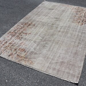 Könnte beinhalten: Ein Vintage-Teppich in gebrochenem Weiß mit einem verblassten Blumenmuster in Braun- und Rosatönen. Der Teppich hat einen ausgefransten Rand und liegt auf einer grauen Asphaltfläche.