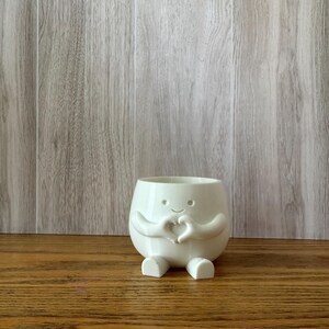 May include: A white ceramic planter shaped like a small, round character with a smiling face. The figure has two arms positioned in front of its chest to form a heart shape with its hands and two small feet extending from the base of the vessel.