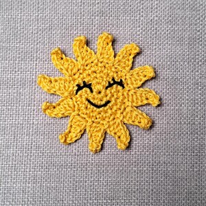 Può includere: Un sole giallo all'uncinetto con un volto sorridente e gli occhi chiusi. Il sole ha più raggi che si estendono verso l'esterno. Il sole è fatto di filato e posto su uno sfondo di tessuto grigio. Perfetto per qualsiasi progetto.