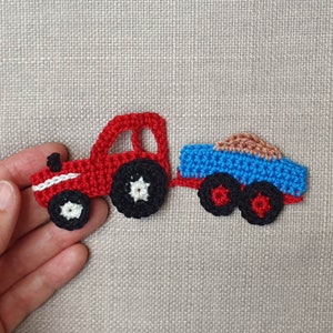 May include: A crocheted tractor in red and black, with a blue trailer carrying brown cargo. The tractor has white detailing and black wheels. The trailer is blue with black wheels and red accents. The item is held against a neutral background.