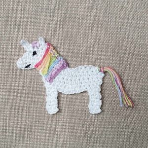 Può includere: Un unicorno bianco all'uncinetto con una criniera e una coda arcobaleno. L'unicorno è in piedi su una superficie marrone testurizzata.