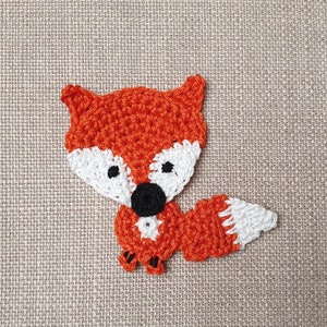 Könnte beinhalten: Ein gehäkelter Fuchs mit orangefarbenem Fell, weißen Akzenten und schwarzen Augen. Der Fuchs sitzt auf einem beigefarbenen Stoffhintergrund.