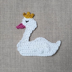 Könnte beinhalten: Ein weißer gehäkelter Schwan mit einem rosa Schnabel und einer goldenen Krone. Der Schwan sitzt auf einem braunen Stoffhintergrund.