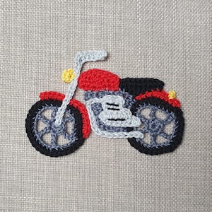 Op de afbeelding: Een gehaakte motorfiets patch met een rode zitting en details, zwarte banden en een grijze carrosserie. Het stuur en de voorvork zijn grijs, met een gele koplamp. De patch staat op een neutraal gekleurde stoffen achtergrond.
