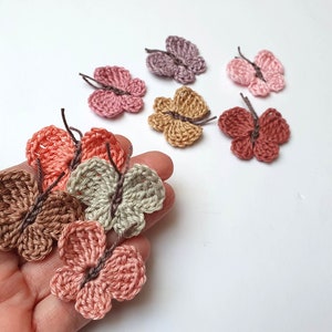 Könnte beinhalten: Eine Sammlung gehäkelter Schmetterlingsverzierungen in verschiedenen Rosatönen, Braun- und Beigetönen. Die Schmetterlinge sind klein und zart mit filigranen Details.