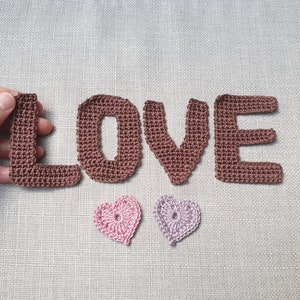 May include: Crocheted brown letters spelling "LOVE" with two small crocheted hearts, one pink and one lavender. The letters and hearts are handmade and placed on a neutral linen background. The letter "L" is being held by a hand.